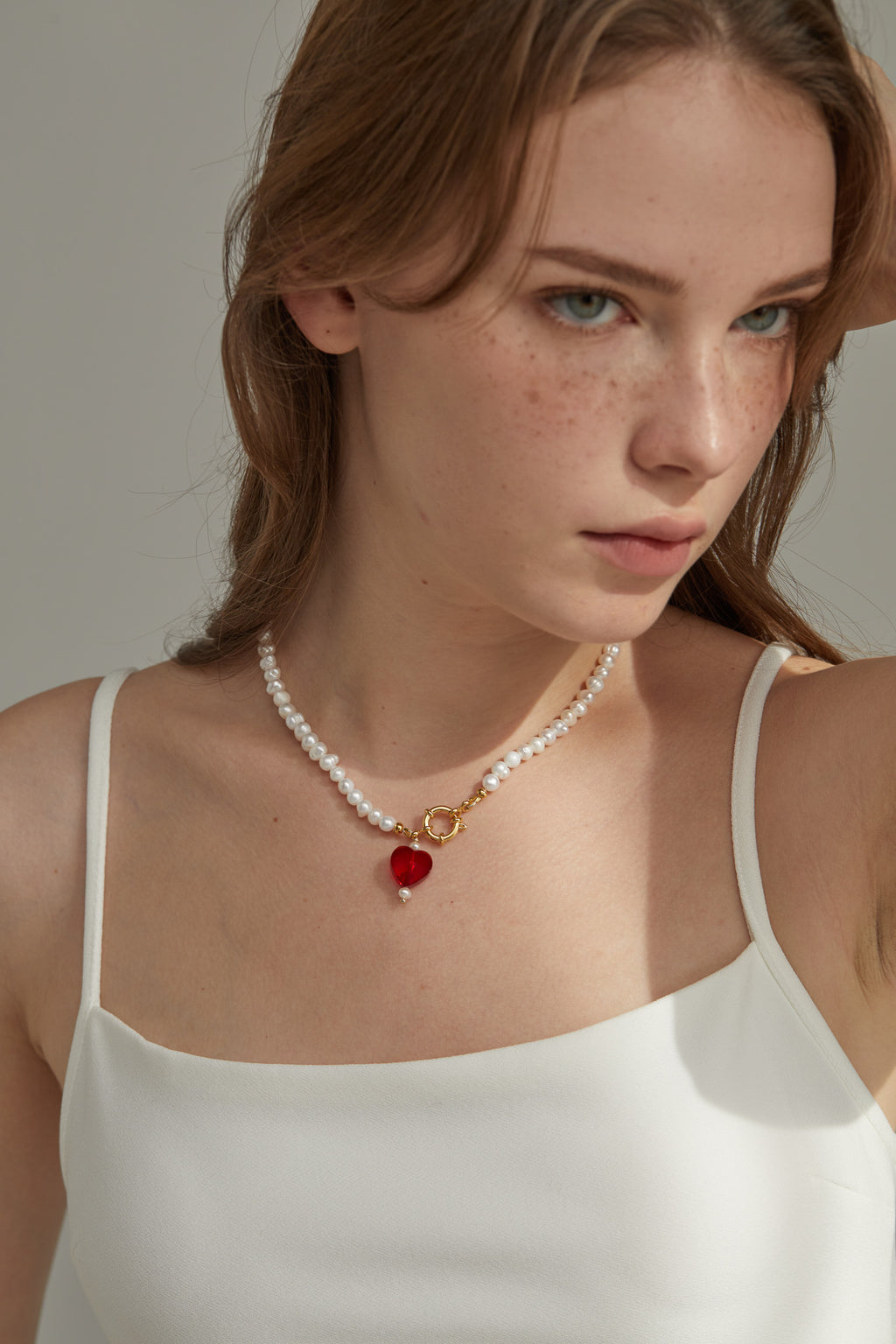 Woman wearing a pearl necklace with a red heart pendant against a neutral background
