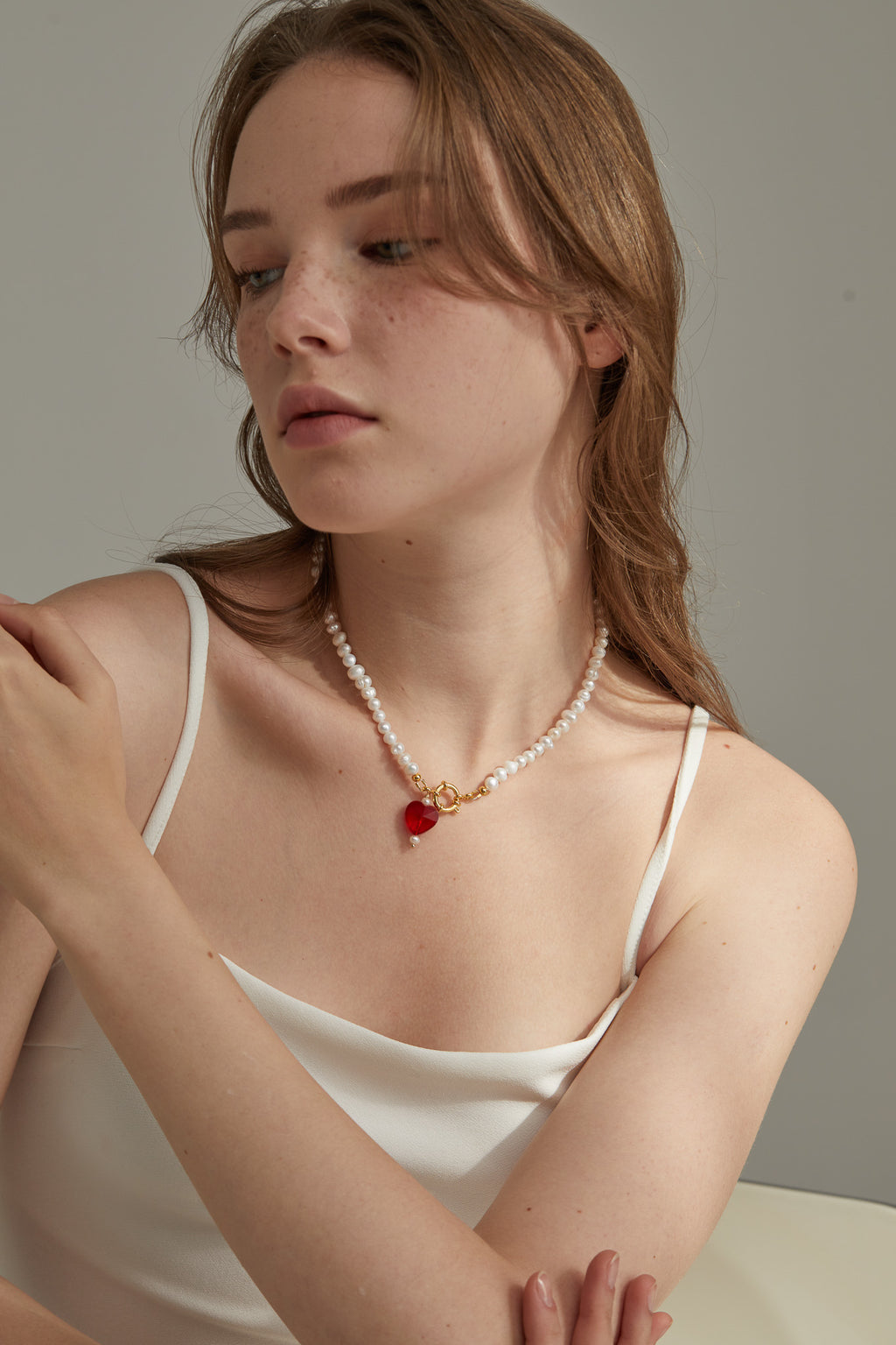 Woman wearing a pearl necklace with a red heart pendant against a neutral background